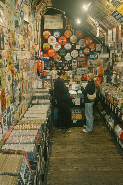 Inside a vintage bookshop, two women are engaged in conversation while examining a selection of books on a central display table. The shop is densely stocked with books along narrow aisles and on book