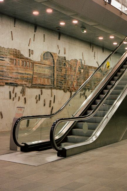 A photograph of a staircase and an escalator inside a building, with the escalator flanked by two sets of traditional stairs on either side. The staircase features carpeted steps with a textured appea