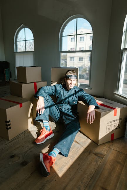 A person sitting on the floor inside a well-lit room with large arched windows, surrounded by several cardboard boxes sealed with red and black packing tape, ready for a home relocation. The individua