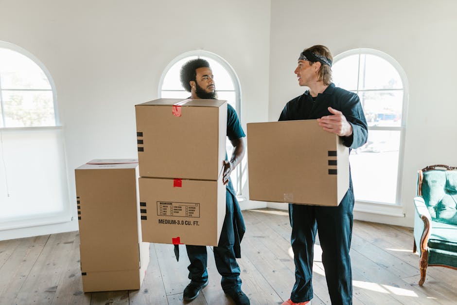 A person sitting on the floor inside a well-lit room with large arched windows, surrounded by several cardboard boxes sealed with red and black packing tape, ready for a home relocation. The individua