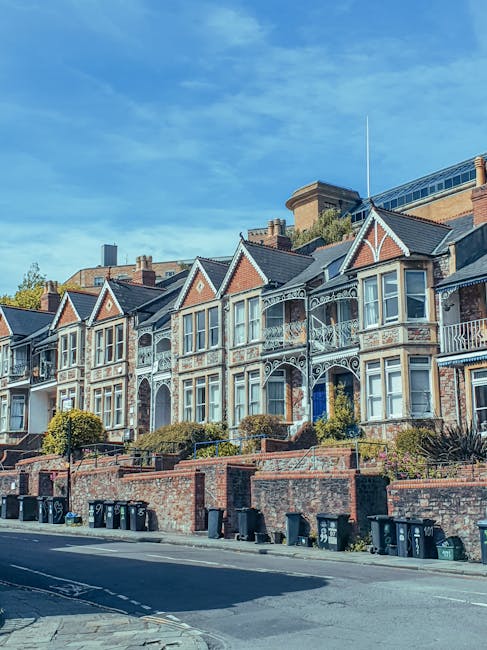 A row of Victorian-style terraced houses with decorative ironwork balconies, pitched roofs with chimney stacks, and large bay windows situated on an elevated street level. The houses are built with re